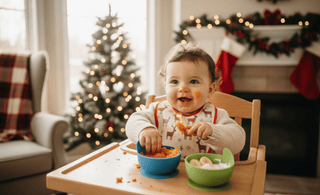 Smiling baby enjoying mashed Christmas baby food in a high chair, wearing a festive reindeer bib, with a decorated Christmas tree and holiday lights in the background — The Mom Store baby feeding essentials.