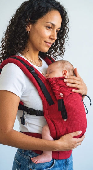 A mother gently cradling her sleeping baby's head, highlighting the folded neck support panel of the red Aruna Yoga baby carrier.