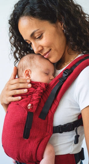 Close-up of a mother resting her cheek against her sleeping infant, both comfortable and secure in the red handwoven cotton Aruna carrier.