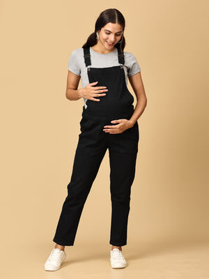 Front view of a Model posing with her hand on pregnant belly in The Mom Store black Denim Maternity Dungaree with Side Buttons 