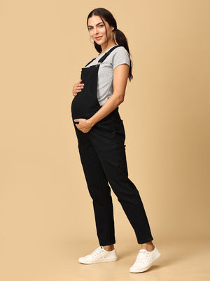 Model posing with her hand on pregnant belly in The Mom Store black Denim Maternity Dungaree with Side Buttons 