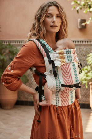 A woman with wavy hair in a terracotta dress wearing the Almitra Tattva Adira Yoga carrier in a front-carry position. The image highlights the carrier's side profile, showing the adjustable black webbing straps, padded shoulder support, and the baby’s ergonomic "M-position" leg placement. The linen fabric displays a striking vertical chevron and diamond motif.