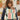 Close-up of a woman wearing the Adira Yoga Baby Carrier in off-white linen with colorful tribal-inspired patterns. The baby is snugly secured in a front inward-facing position. The background features rustic terracotta pots and a wooden trellis, emphasizing the brand's earthy, sustainable, and inclusive aesthetic.