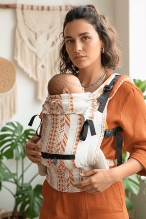 Woman using Almitra Tattva ergonomic linen baby carrier in rust and natural tones, newborn baby secured in front carry position, adjustable straps, inclusive babywearing design, breathable fabric