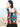 A woman with a shoulder tattoo wearing a vibrant Almitra Tattva woven baby carrier in a back carry position. The carrier features a bold, high contrast geometric pattern in turquoise, magenta, black, and orange. A toddler is seated ergonomically inside, looking over the wearer's shoulder. The image is set outdoors with soft natural lighting, emphasizing the rich texture.