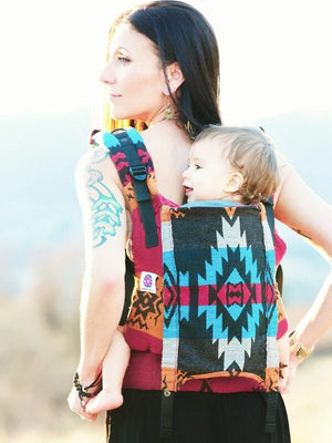 A woman with a shoulder tattoo wearing a vibrant Almitra Tattva woven baby carrier in a back carry position. The carrier features a bold, high contrast geometric pattern in turquoise, magenta, black, and orange. A toddler is seated ergonomically inside, looking over the wearer's shoulder. The image is set outdoors with soft natural lighting, emphasizing the rich texture.