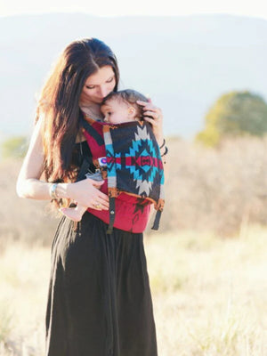 Mother holding baby close in Almitra Tattva Kullu-inspired ergonomic baby carrier featuring bold tribal weave patterns in red, turquoise, black, and ochre tones, designed for inclusive babywearing, hands-free comfort, and secure front carry support outdoors.