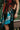 A mother wearing a newborn in an Almitra Tattva Auronee woven wrap featuring vibrant turquoise, rust orange, and black geometric tribal patterns. The baby is held securely in a front-facing ergonomic carry against the mother’s chest.