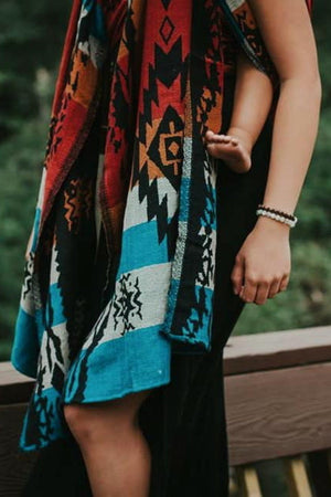 A mother wearing a newborn in an Almitra Tattva Auronee woven wrap featuring vibrant turquoise, rust orange, and black geometric tribal patterns. The baby is held securely in a front-facing ergonomic carry against the mother’s chest.