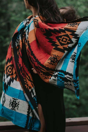 Side view detail of the Almitra Tattva Auronee wrap draped over a caregiver's shoulder, highlighting the intricate black diamond and chevron motifs on a background of turquoise, orange, and white bands. A baby's foot is visible, showing the wrap's supportive texture.