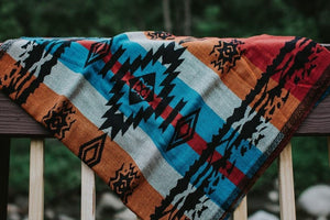 The Almitra Tattva Auronee woven wrap displayed flat over a wooden railing, showcasing the full geometric tribal design, featuring large black diamond centerpieces flanked by colorful turquoise and burnt orange stripes.
