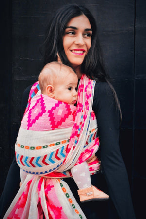 Mother carrying baby in Almitra Tattva Shubra woven baby wrap featuring bright pink geometric motifs and multicolor bohemian woven borders outdoors.