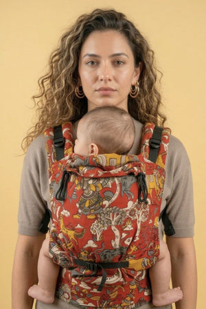 Parent wearing the Almitra Tattva Jataka Tales Kalamkari baby carrier in a front-carry position, featuring bold handcrafted-inspired Indian folk art prints in earthy red and mustard tones. Ergonomic soft structured baby carrier designed for newborns, infants, and toddlers with adjustable padded straps for inclusive, comfortable babywearing.