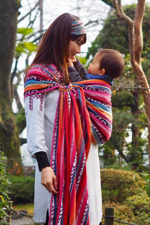 Full length image of caregiver using Almitra Tattva Larona Wrap Converted Ring Sling baby carrier outdoors, showcasing colorful woven boho design, comfortable hip carry position, and inclusive ergonomic support.
