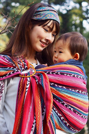 Mother carrying baby in Almitra Tattva Larona Wrap Converted Ring Sling featuring vibrant bohemian woven fabric with ergonomic babywearing support, inclusive hands free baby carrier for newborns and toddlers.