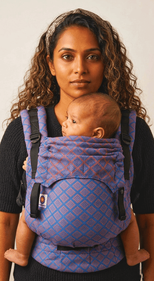 Front portrait of a woman wearing the Almitra Tattva Nilopala carrier, displaying the full front body panel, folded neck support, and wide patterned waistband.