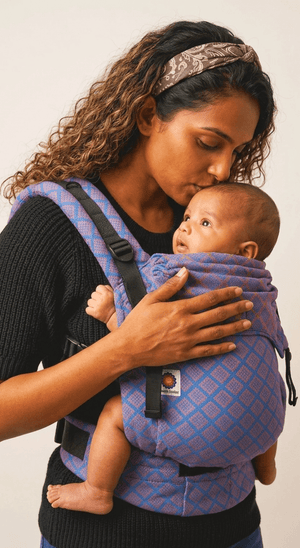 Close-up of a mother gently kissing the forehead of her sleeping baby secured in the blue patterned Nilopala baby carrier.