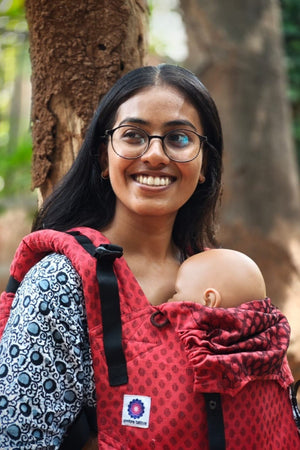 Young mother outdoors using Almitra Tattva red sustainable baby carrier with ergonomic shoulder support, breathable fabric, and comfortable front babywearing design.