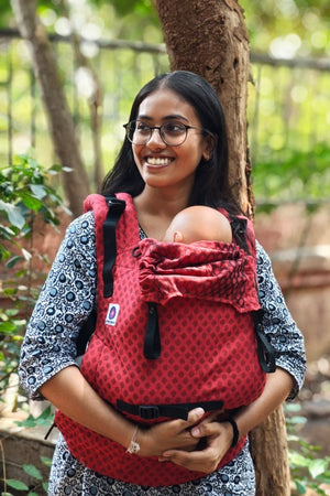 Smiling mother wearing Almitra Tattva red ergonomic baby carrier outdoors, carrying baby safely in a sustainable cotton babywearing carrier with adjustable straps and inclusive comfort design.