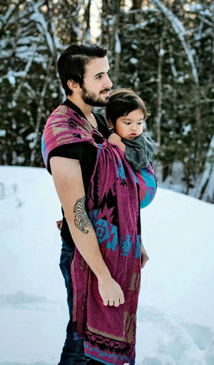 Side-view of a father using a handwoven Almitra Tattva Pema Ring Sling to carry a child in a snowy forest. The image showcases the intricate Kullu weave and the supportive, ergonomic fit of the fabric across the wearer's shoulder and the baby's back.