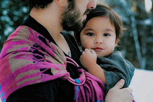 Full-length lifestyle shot of a man wearing a toddler in a pink and blue Almitra Tattva Pema Ring Sling against a snowy backdrop. The image illustrates the long tail of the handwoven wrap and its stylish, gender-neutral appeal for modern parents.