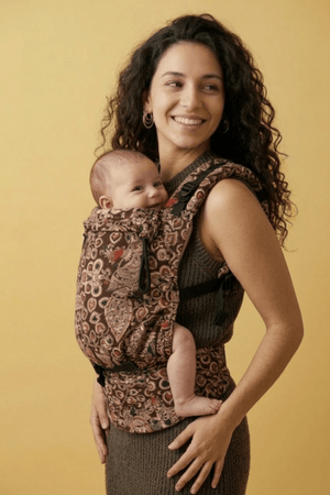 Smiling mother and baby in Almitra Tattva paisley soft structured carrier with ergonomic front carry position, featuring earthy neutral aesthetic, adjustable waist support, inclusive babywearing design, breathable woven fabric, and cozy everyday comfort for infants and caregivers.