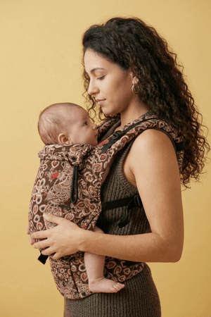 Mother carrying newborn in Almitra Tattva paisley print soft structured baby carrier in ergonomic front carry position, featuring earthy brown tones, adjustable straps, inclusive babywearing design, and breathable woven fabric for newborn comfort and parent support.