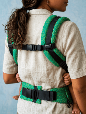 Rear view of a woman showing the horizontal chest clip, padded green shoulder straps, and wide lumbar waist buckle of the carrier.