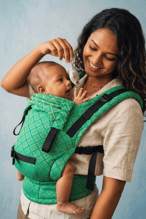 Mother holding a small toy above her alert baby seated in the green handwoven Mangalgiri cotton Yaman Yoga carrier.