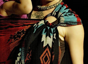 A close-up, high-contrast shot of the Almitra Tattva Auronee wrap fabric used in a secure back carry. The image highlights the intricate weave and vibrant earth-toned geometric motifs against a dark background.