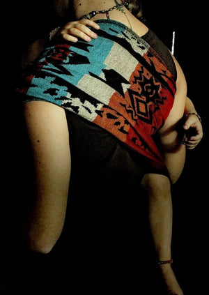 A mother wearing a toddler in the Almitra Tattva Auronee ring sling from the Kullu Collection. The handwoven fabric features bold geometric patterns in rust red, turquoise, and black, styled with a matching headwrap for an inclusive, artistic look.