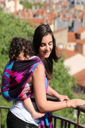 A mother smiling while using the Almitra Tattva Pema woven wrap from the Kullu Collection for a secure back carry. The wrap features a bold geometric pattern in magenta, turquoise, and black.