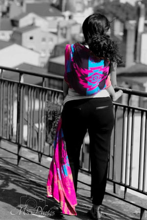 Full-length view of a caregiver wearing the Almitra Tattva Pema wrap in a back carry position against a city backdrop, highlighting the long, flowing fabric tails and inclusive sizing.