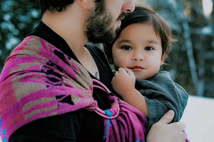 Full-length lifestyle shot of a man wearing a toddler in a pink and blue Almitra Tattva Pema Ring Sling against a snowy backdrop. The image illustrates the long tail of the handwoven wrap and its stylish, gender-neutral appeal for modern parents.