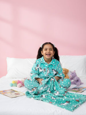 Child wearing teal pajamas with unicorn prints, sitting on a bed against a pink wall