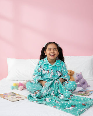 Child wearing teal pajamas with unicorn prints, sitting on a bed against a pink wall