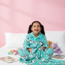 Child wearing teal pajamas with unicorn prints, sitting on a bed against a pink wall