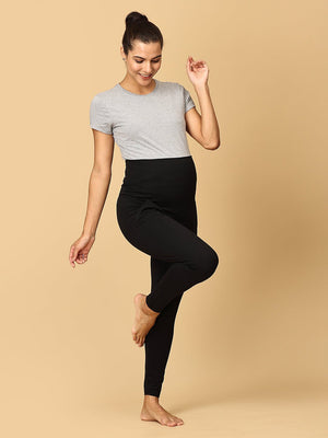 A pregnant woman wearing black maternity leggings with a wide, belly support waistband, paired with a short grey top.
