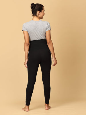 A pregnant woman wearing black maternity leggings with a wide, belly support waistband, paired with a short grey top.