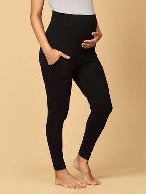 A pregnant woman wearing black maternity leggings with a wide, belly support waistband, paired with a short grey top.