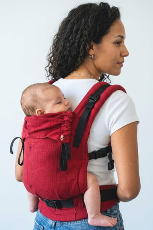 A baby looking over their mother's shoulder while seated safely in the red Almitra Tattva Aruna handwoven cotton baby carrier.