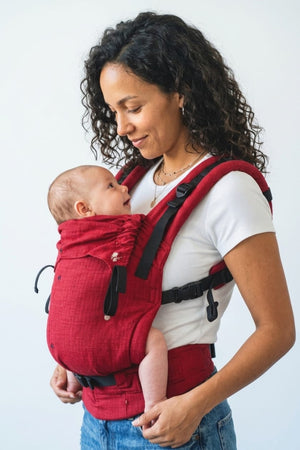 Side profile of a mother and baby making eye contact, showcasing the ergonomic seat and padded shoulder straps of the red Aruna cotton carrier.