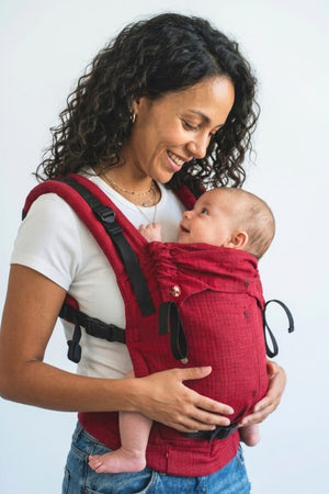 Front view of a smiling mother wearing the solid crimson red Almitra Tattva Aruna full-buckle baby carrier, securely holding her infant.
