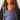 Front portrait of a woman wearing the Almitra Tattva Nilopala carrier, displaying the full front body panel, folded neck support, and wide patterned waistband.