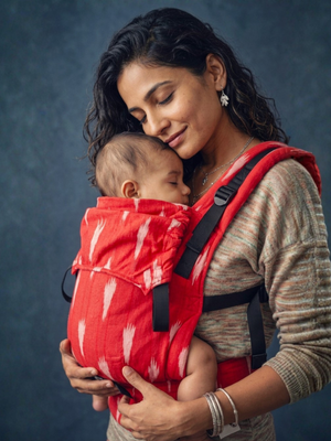 A mother gently resting her cheek against her sleeping baby's head in the red handwoven cotton Shendur Ikkat carrier.