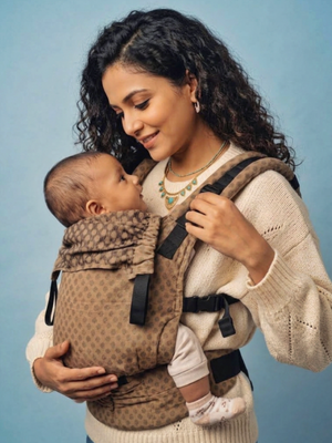 Mother smiling at her alert baby inside the mocha-patterned Almitra Tattva Tushi carrier, showing the padded shoulder straps and side buckles.