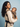 Side angle of a woman standing with her baby resting inward in the Tushi carrier, displaying the supportive waistband and black safety webbing.