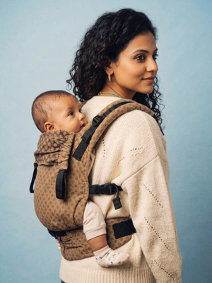 Rear-side view of a mother carrying her baby on her front, showing the baby looking back over her shoulder and the ergonomic strap structure.