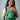 Front view of a smiling mother standing and wearing the green diamond-patterned Almitra Tattva Yaman Yoga carrier with her baby positioned inside.