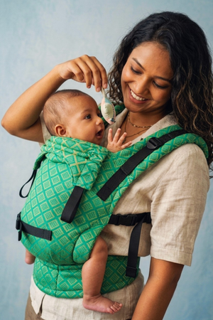 Mother holding a small toy above her alert baby seated in the green handwoven Mangalgiri cotton Yaman Yoga carrier.